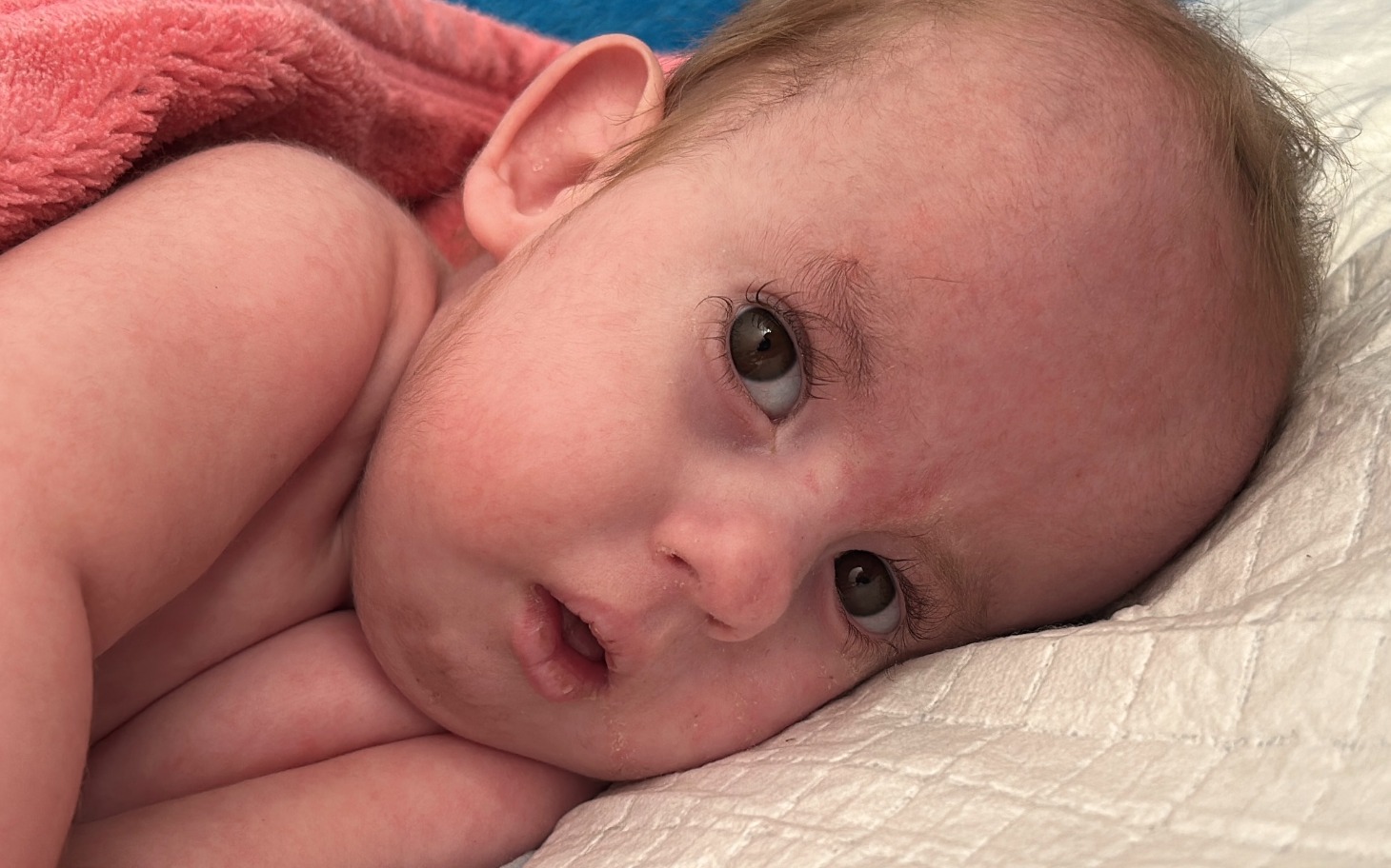 
        Małe dziecko leżące na poduszce, z wyrazem twarzy pełnym ciekawości.
    