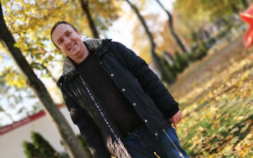 
        Uśmiechnięty mężczyzna w parku, otoczony jesiennymi liśćmi.
    