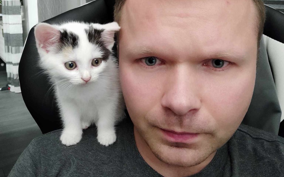 
        Mężczyzna z kotem na ramieniu, obaj patrzą w obiektyw.
    