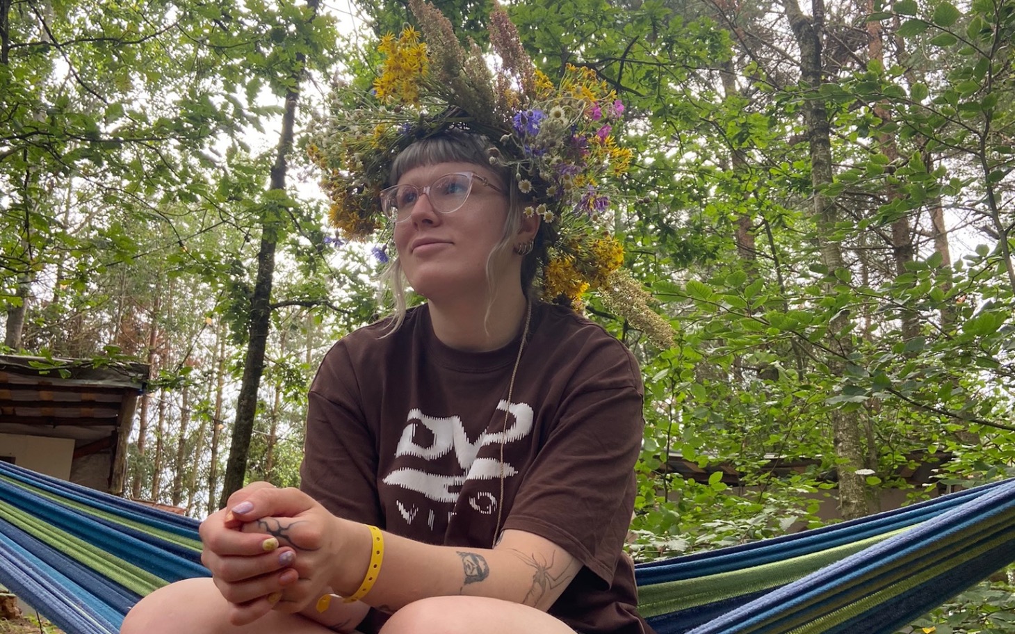 
        Osoba w lesie z kwiatową koroną, relaksująca się na hamaku.
    