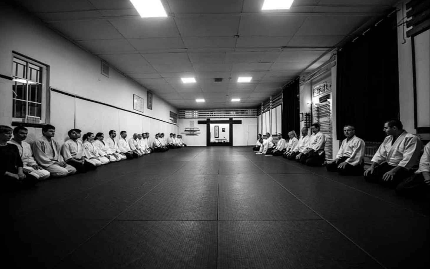 
        Grupa osób w białych giwerach siedzi w półkolu na macie treningowej w dojo
    