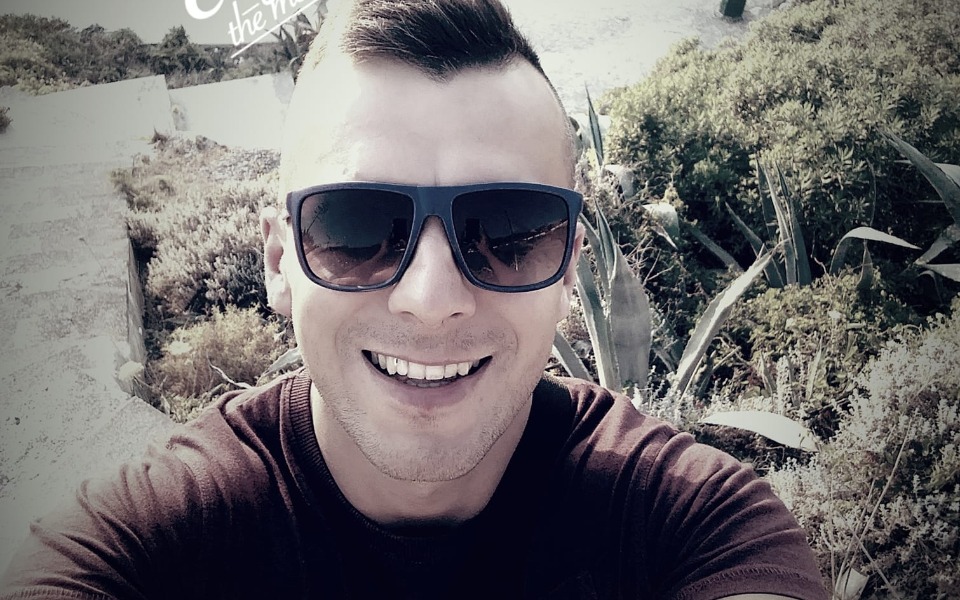 
        Mężczyzna uśmiechający się na tle zieleni, w okularach przeciwsłonecznych.
    