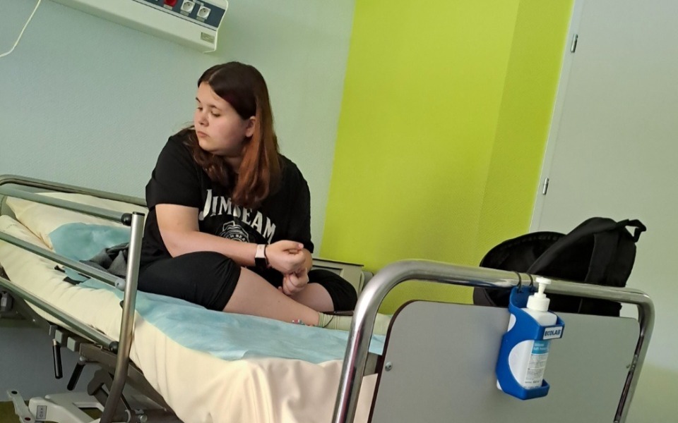 
        Młoda dziewczyna siedzi na łóżku szpitalnym, zamyślona.
    