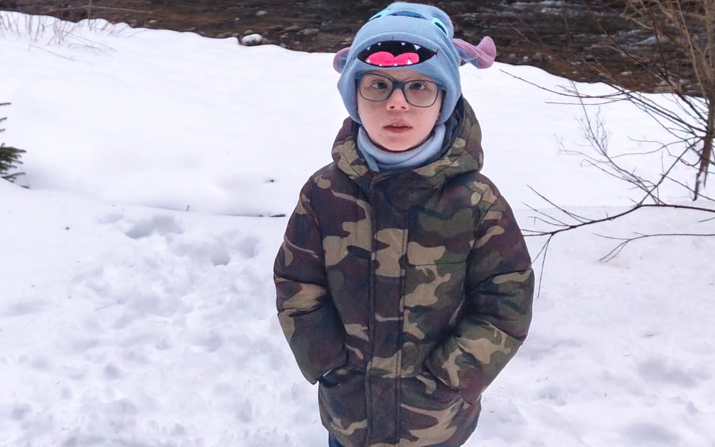 
        Chłopiec w kamuflażowej kurtce i niebieskiej czapce na tle zimowego krajobrazu.
    