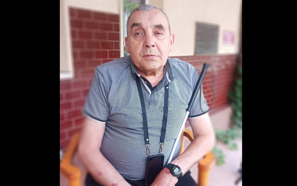 
        Mężczyzna siedzący na krześle, z białą laską.
    