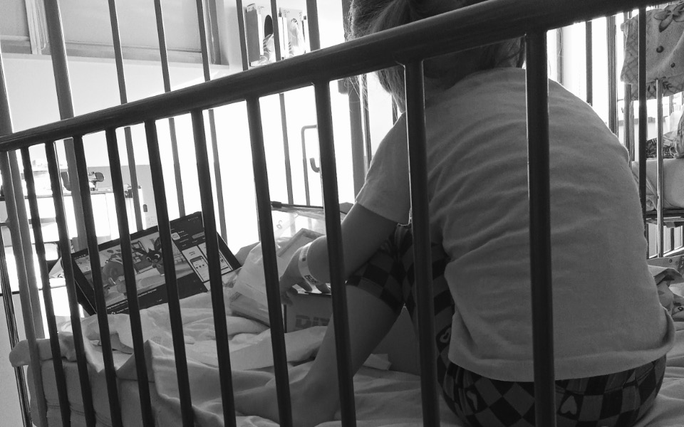 
        Dziecko siedzące w łóżku szpitalnym, oglądające coś na ekranie.
    