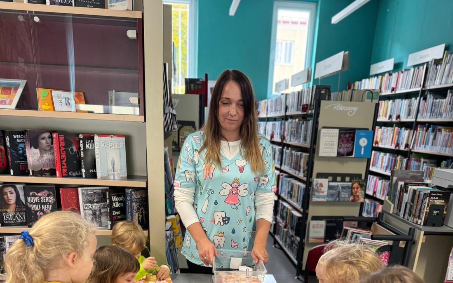 
        Kobieta w bibliotece z dziećmi, prowadząca zajęcia z książkami.
    