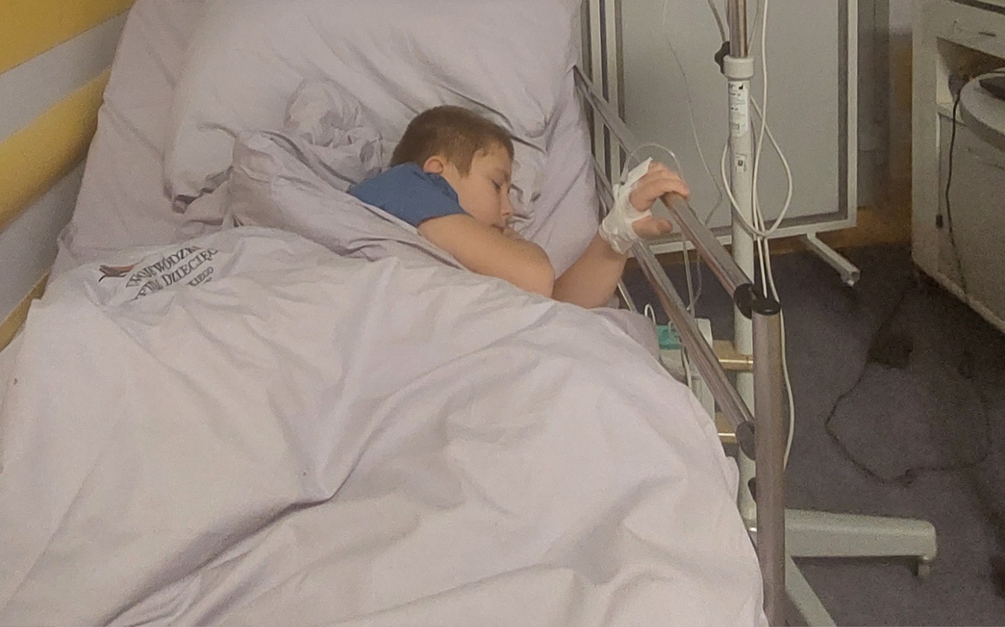 
        Chłopiec śpiący w łóżku szpitalnym, z ręką na barierce.
    