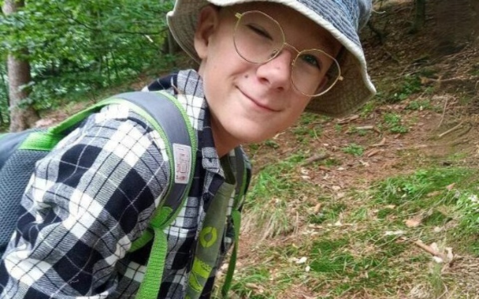 
        Chłopiec w kapeluszu na tle lasu, uśmiechający się do kamery.
    