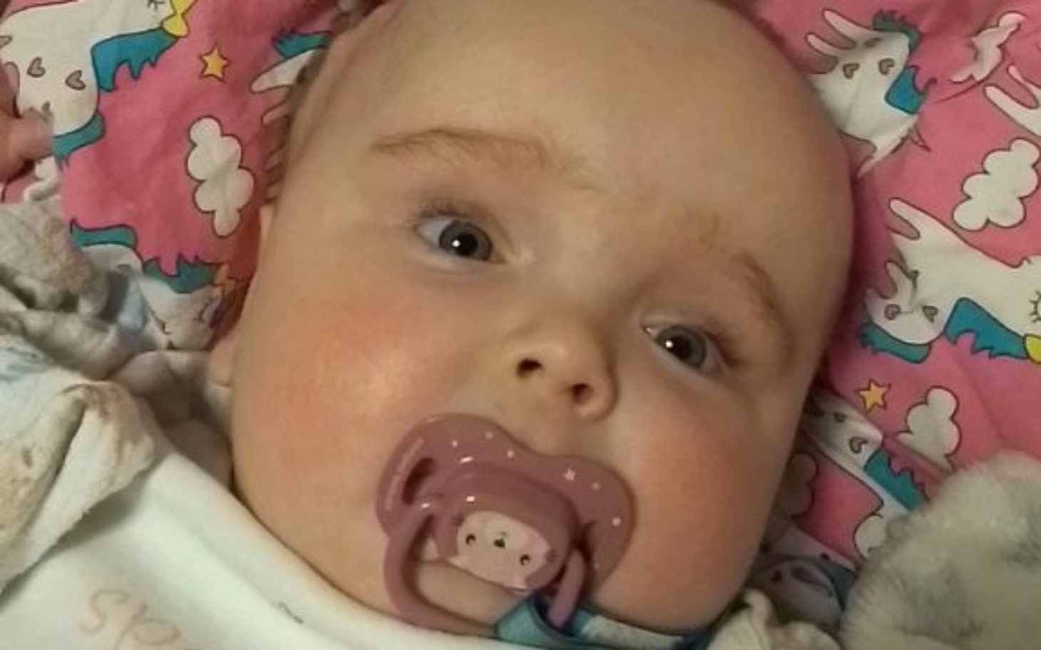 
        Małe dziecko z smoczkiem, leżące na kolorowej pościeli.
    