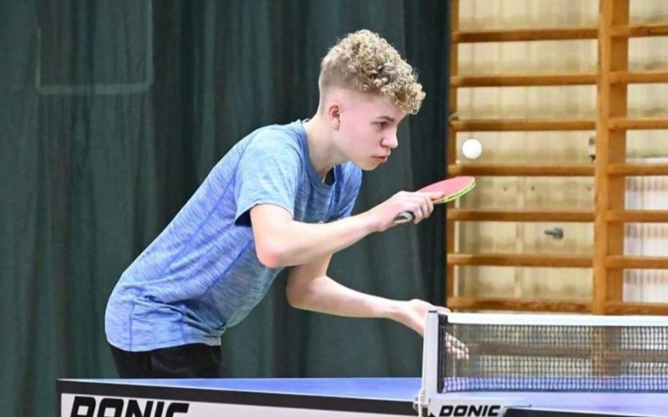 
        Młody chłopak grający w tenisa stołowego, skupiony na grze.
    