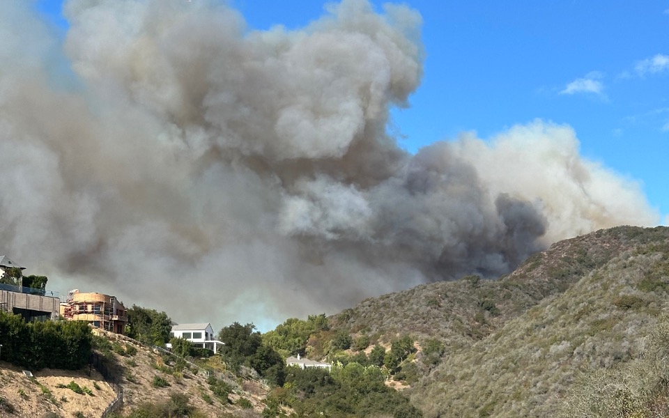 
        Widok na dym unoszący się nad wzgórzami, w tle domy.
    