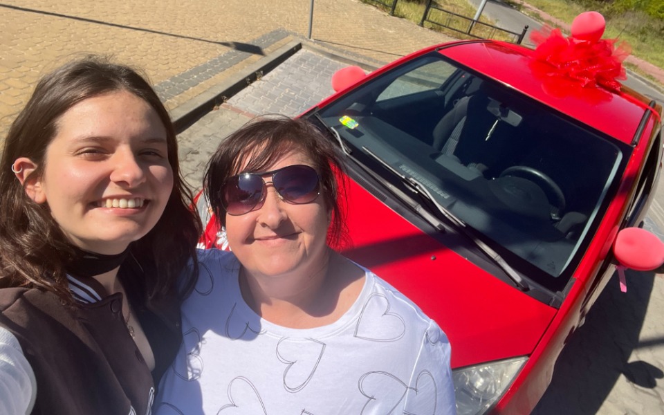 
        Dwie kobiety pozują do zdjęcia obok czerwonego samochodu ozdobionego balonami.
    