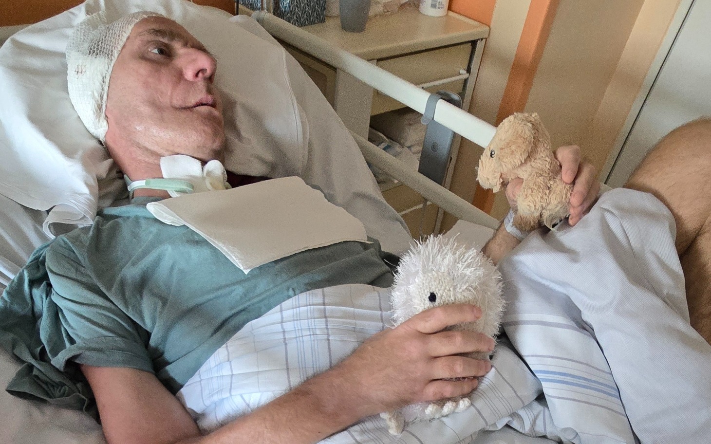 Mężczyzna w szpitalu trzymający pluszowe zabawki, z opatrunkiem na głowie.