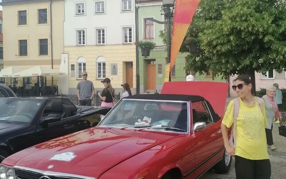 
        Czerwony kabriolet na rynku, w tle ludzie i kolorowe budynki.
    