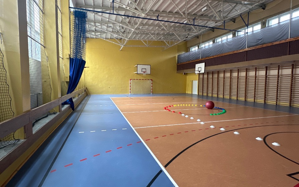 
        Pusta sala sportowa z piłką i kolorowymi znacznikami na podłodze.
    