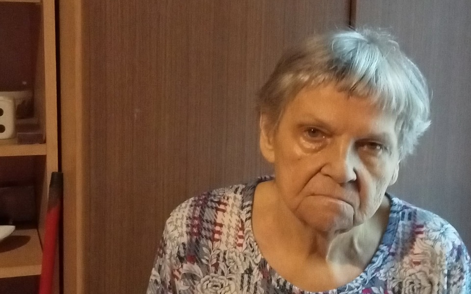 
        Starsza kobieta z poważnym wyrazem twarzy w domowym otoczeniu.
    