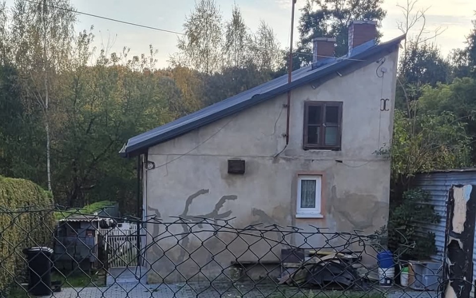 
        Dom z dachem w kształcie spadowym, otoczony zielenią.
    