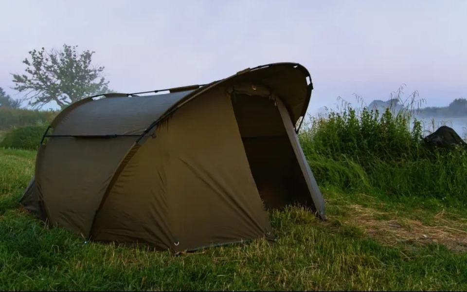 
        Namiot rozstawiony na łące, otoczony mgłą i zielenią.
    