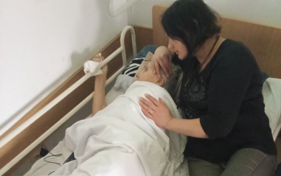 
        Kobieta pociesza starszą osobę leżącą w łóżku.
    