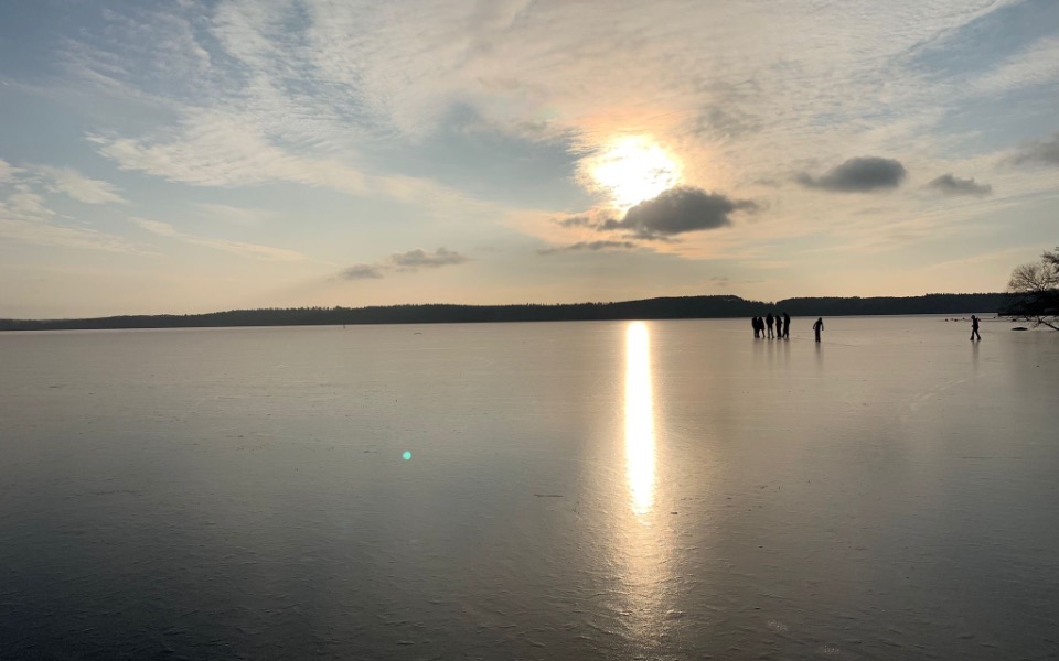
        Widok na zamarznięte jezioro z ludźmi na tle zachodzącego słońca.
    