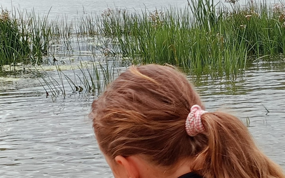 
        Kobieta z długimi włosami nad wodą, w tle roślinność.
    