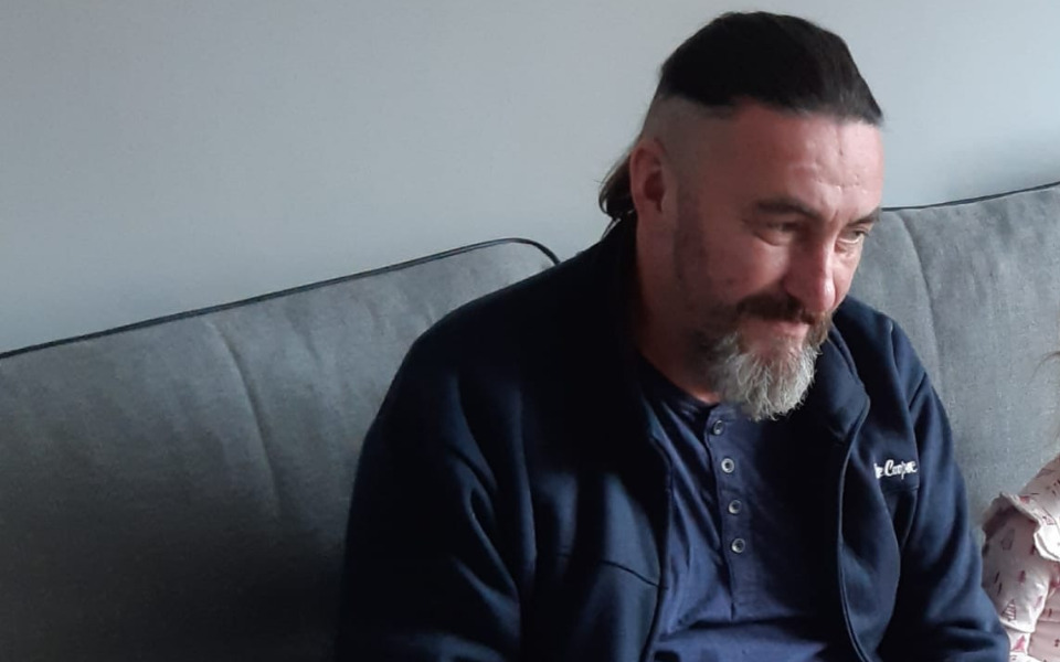 Mężczyzna w szarej bluzie siedzi na kanapie, uśmiechając się.