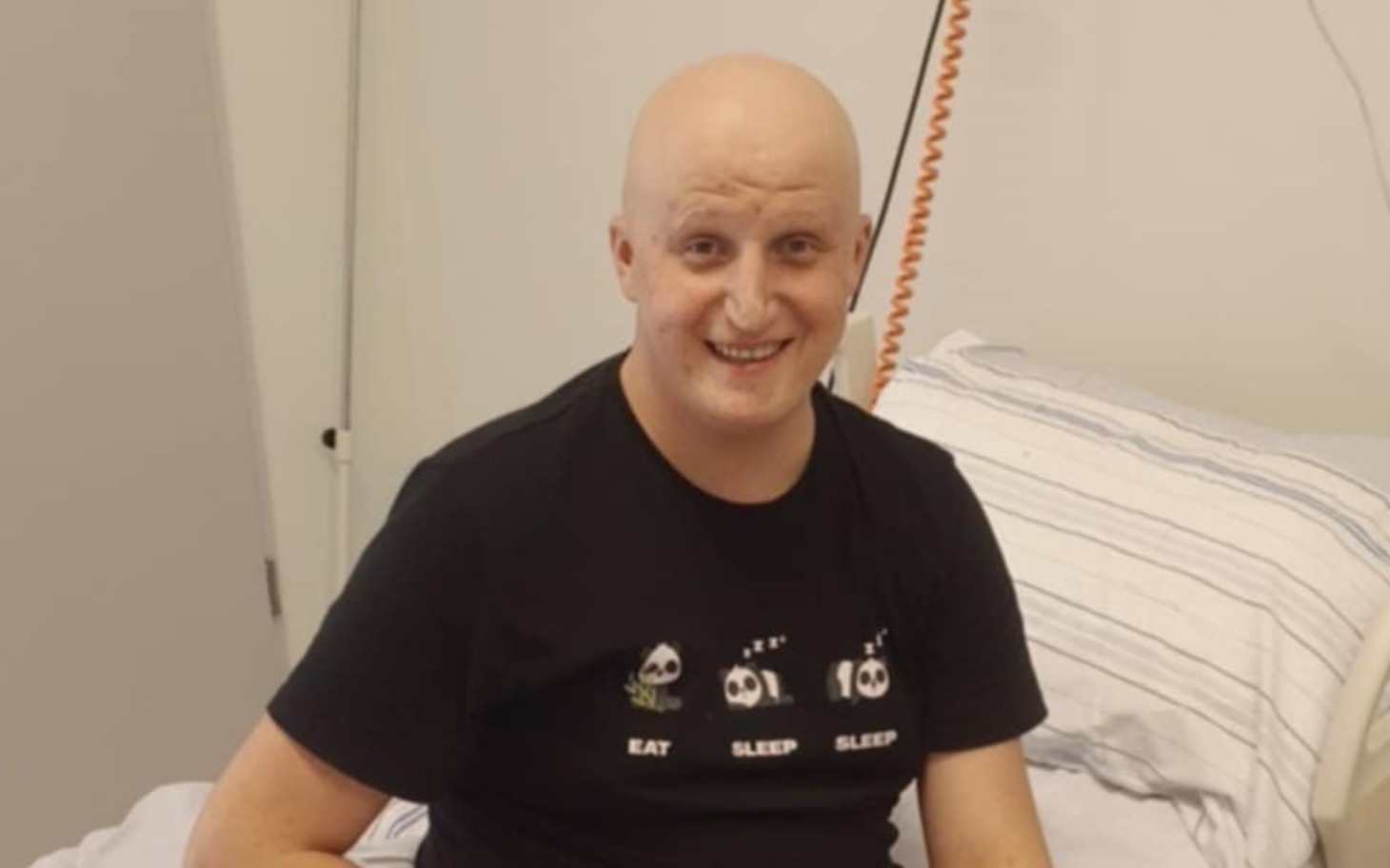 
        Mężczyzna w koszulce z napisem, uśmiechający się w szpitalnym łóżku.
    