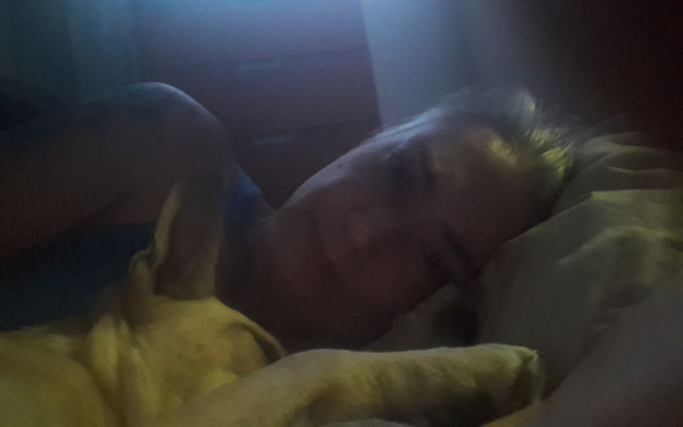 
        Kobieta leżąca w łóżku z psem, w słabym świetle.
    