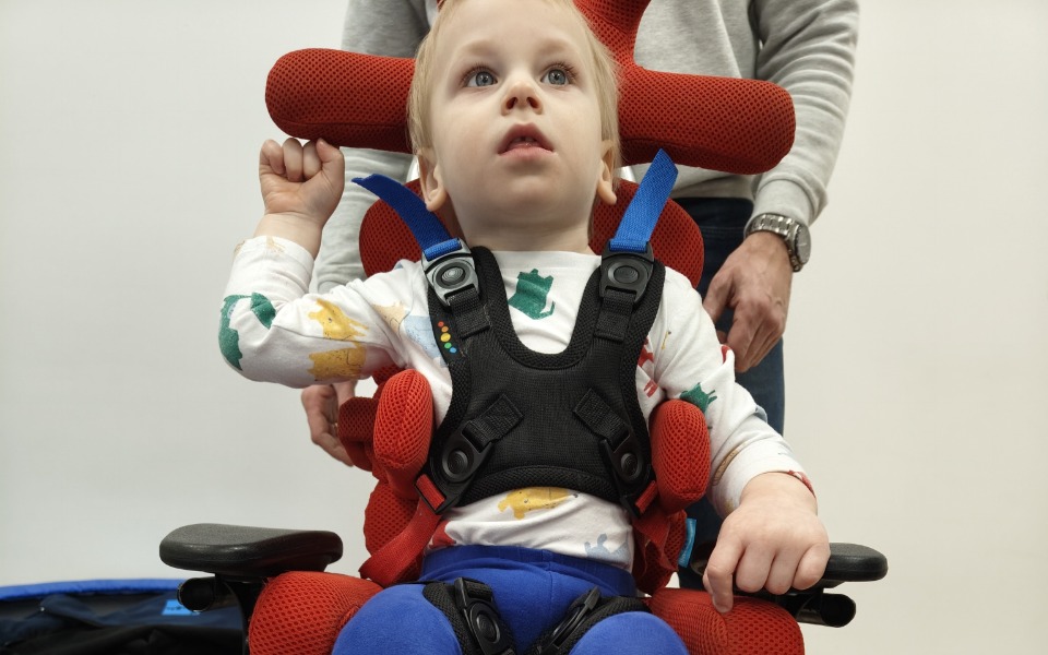 
        Dziecko w specjalnym foteliku, z uśmiechem na twarzy.
    