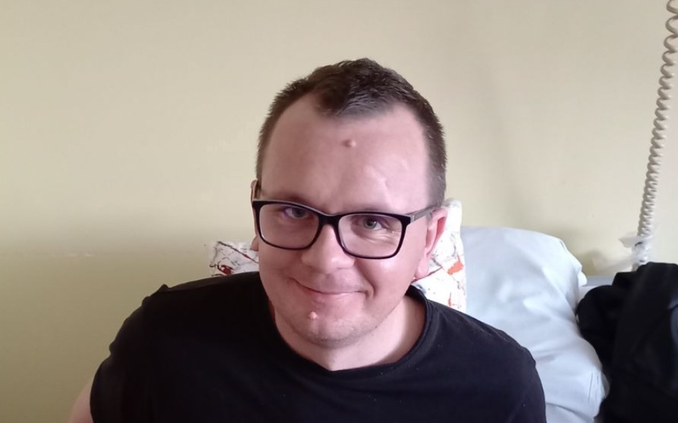 
        Mężczyzna w okularach siedzący na łóżku, uśmiechający się.
    