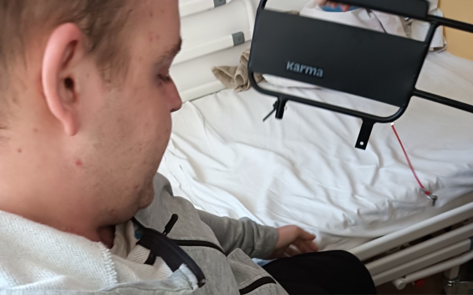 
        Mężczyzna siedzący na łóżku, z ręką na pościeli.
    