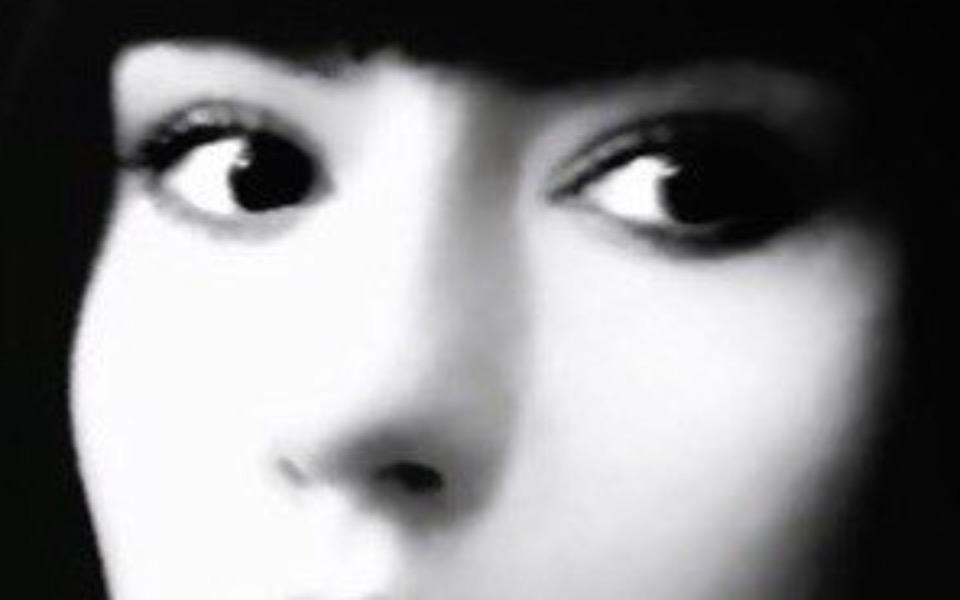 
        Czarno-białe zbliżenie na twarz, z wyraźnymi oczami i tajemniczym wyrazem.
    