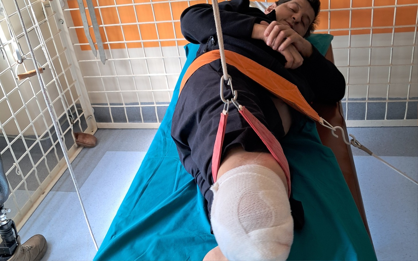 
        Mężczyzna leżący na łóżku szpitalnym z unieruchomioną nogą.
    