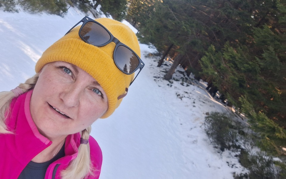 
        Kobieta w żółtej czapce na tle zaśnieżonego lasu.
    