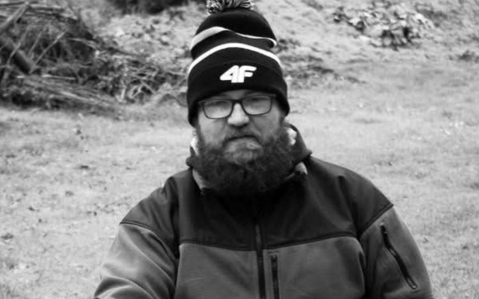 
        Mężczyzna z brodą w czapce, siedzący na trawie.
    