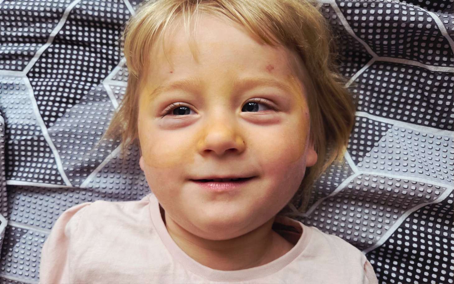 
        Mała dziewczynka z uśmiechem leży na szarym kocyku.
    