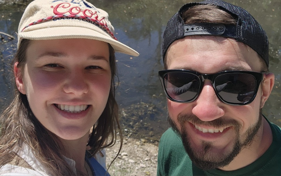 
        Młoda para uśmiechająca się do zdjęcia nad wodą w słoneczny dzień.
    