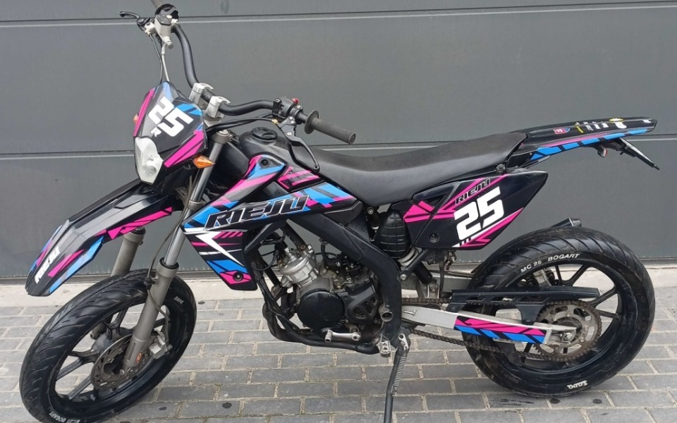 
        Motocykl w kolorach różowym i niebieskim, zaparkowany przy ścianie.
    