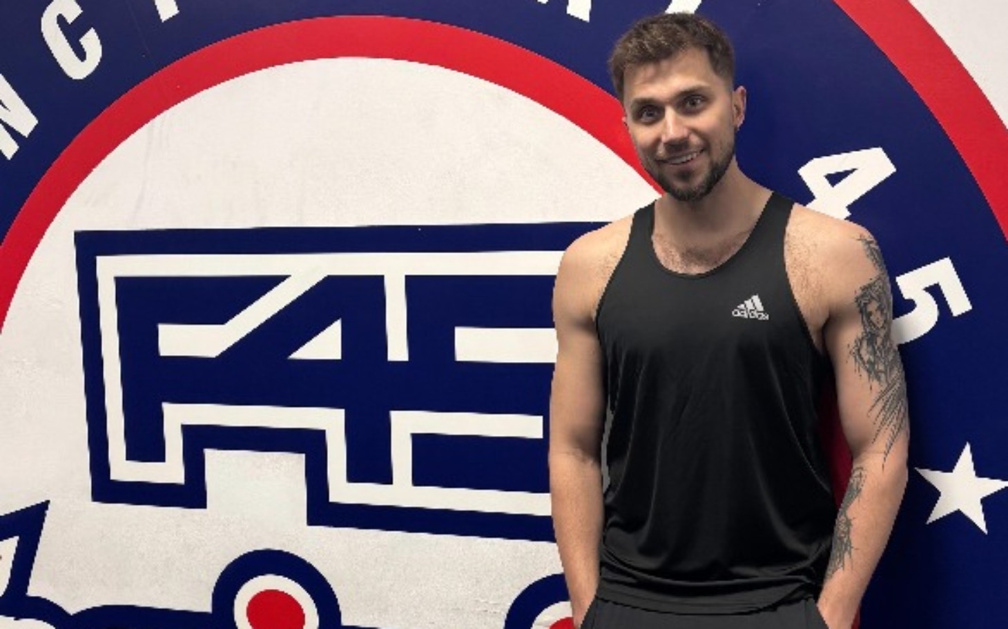 
        Mężczyzna w czarnej koszulce sportowej przy logo klubu fitness.
    