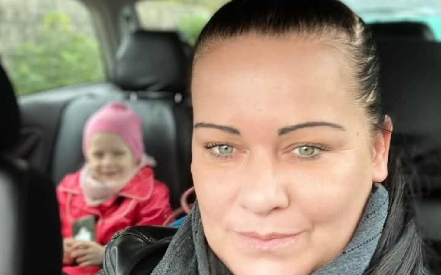 
        Kobieta w samochodzie, z dzieckiem w tle, obie uśmiechnięte.
    