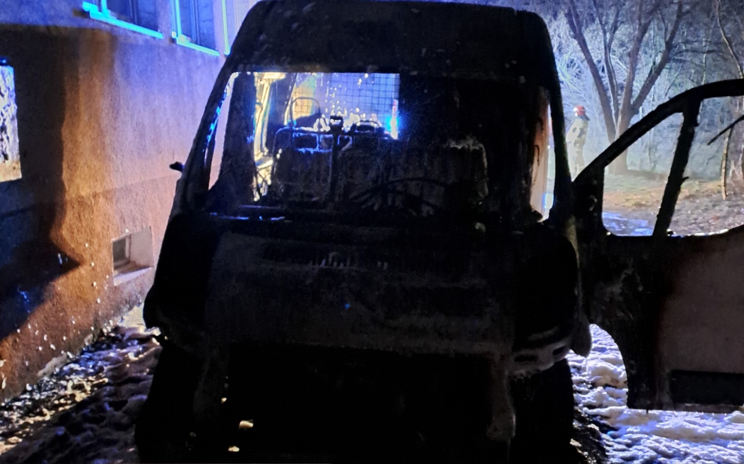 
        Spalony pojazd zaparkowany przy budynku, w tle widać strażaków.
    