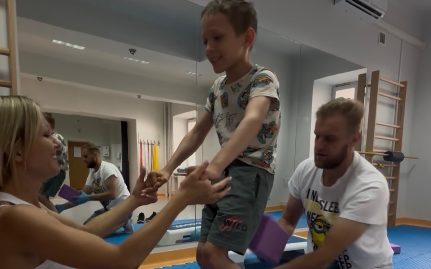 
        Chłopiec ćwiczy z pomocą dorosłych w sali gimnastycznej.
    