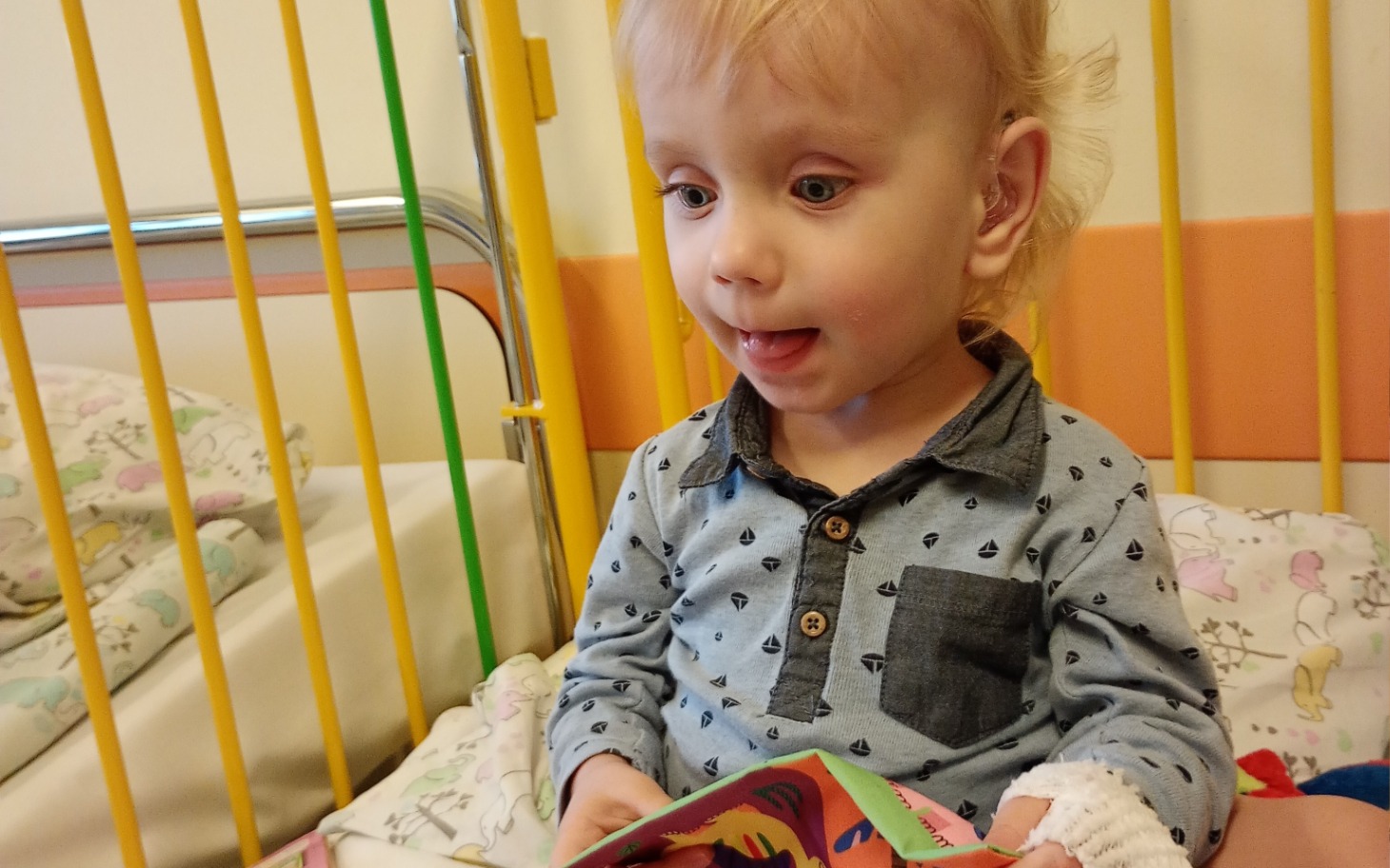 
        Małe dziecko bawiące się książką w szpitalnym łóżku.
    