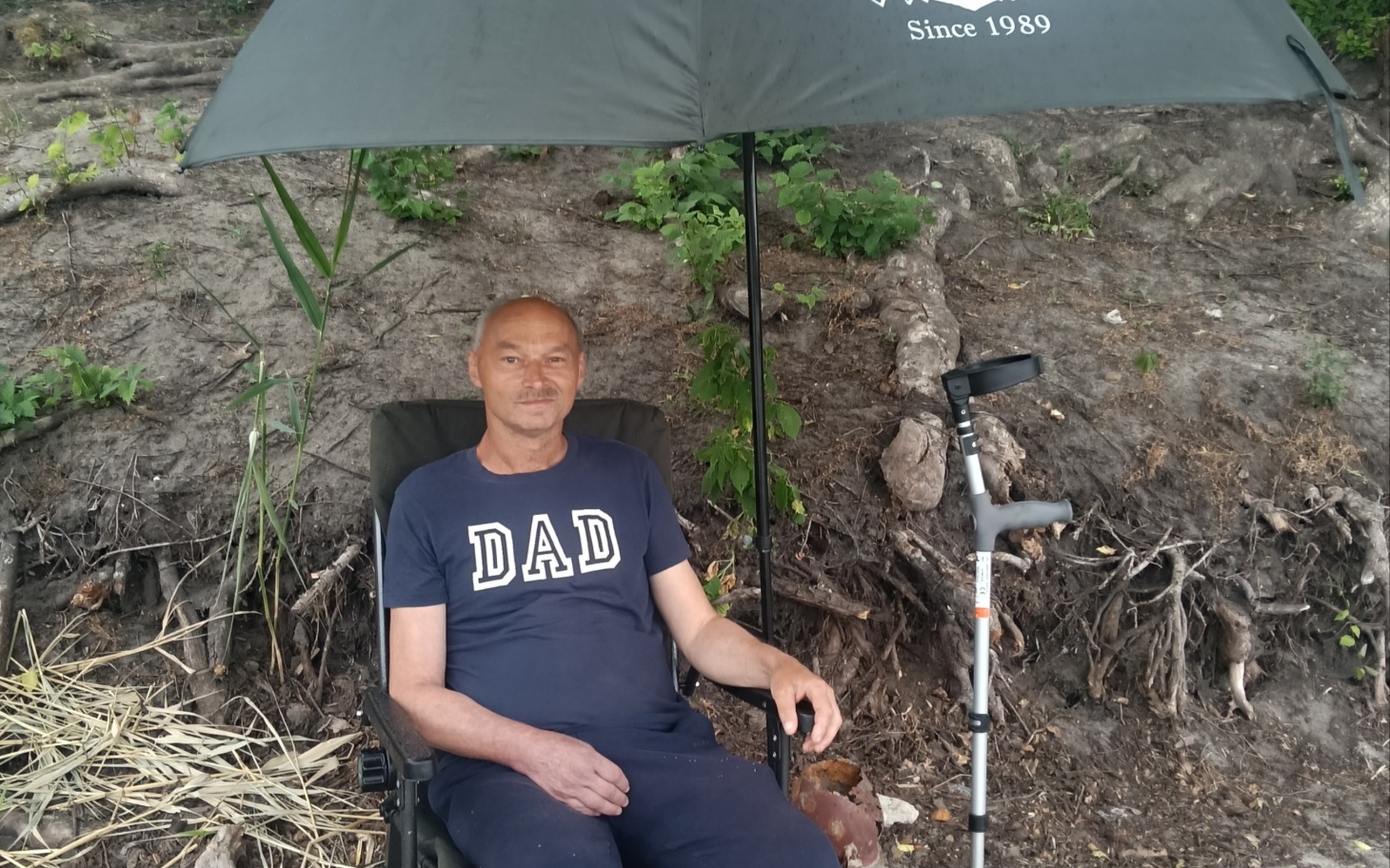 
        Mężczyzna siedzi w krześle pod parasolem w plenerze, uśmiecha się spokojnie.
    