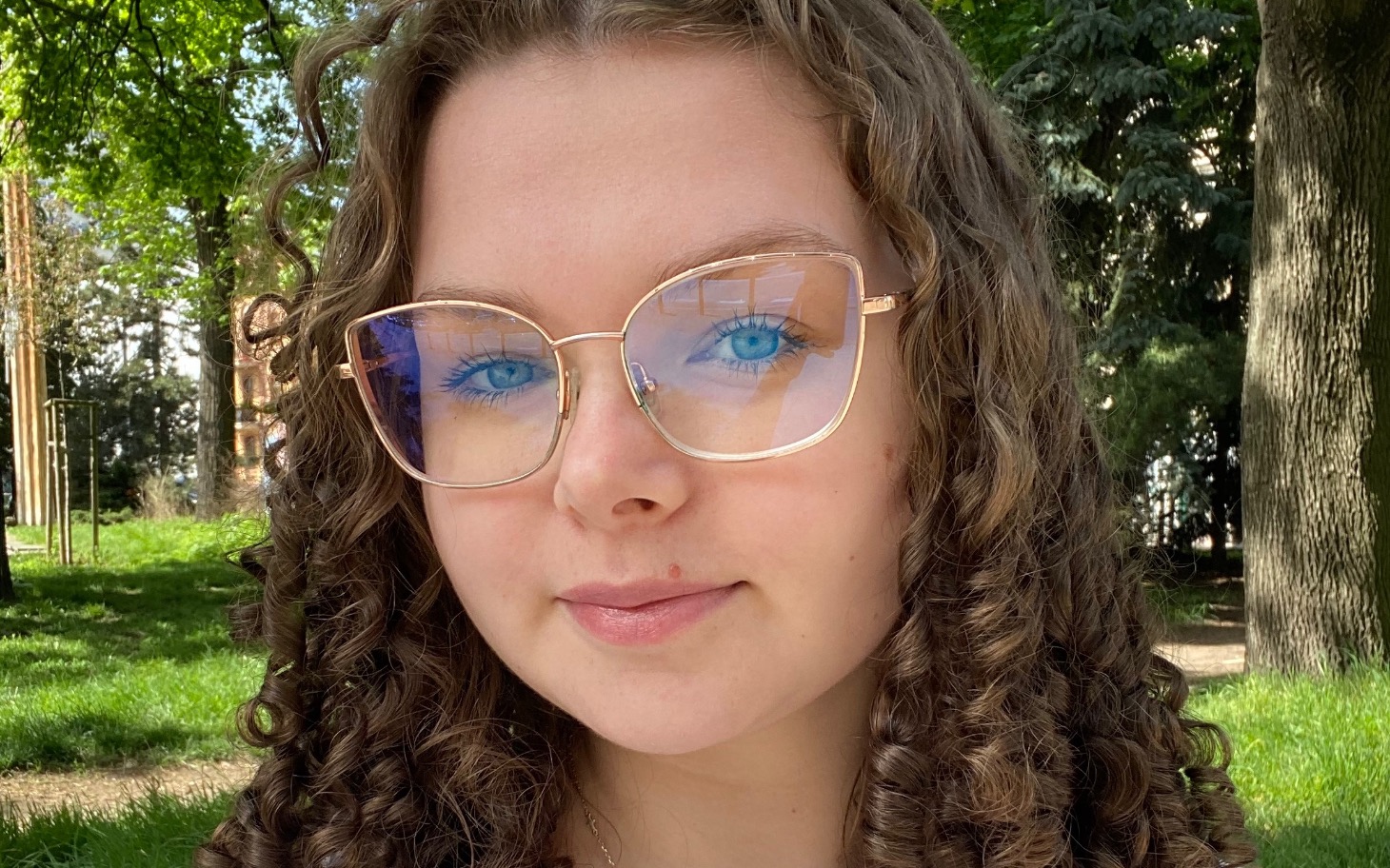 
        Młoda kobieta z kręconymi włosami, w okularach, na tle zieleni.
    