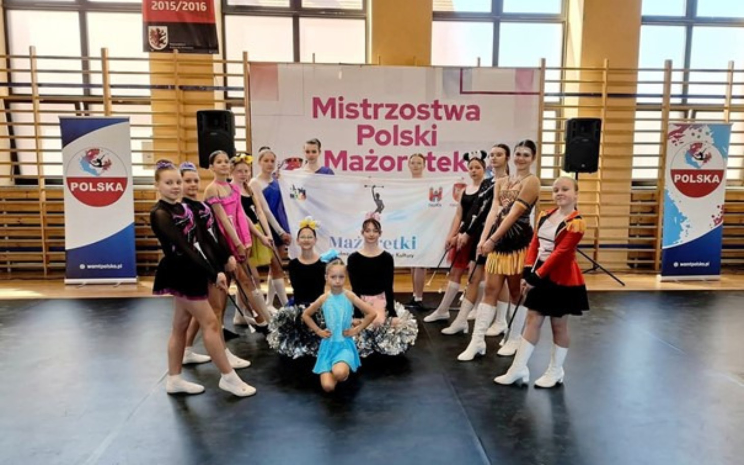 
        Zespół mażoretek na mistrzostwach Polski, uśmiechnięte dziewczyny w strojach.
    