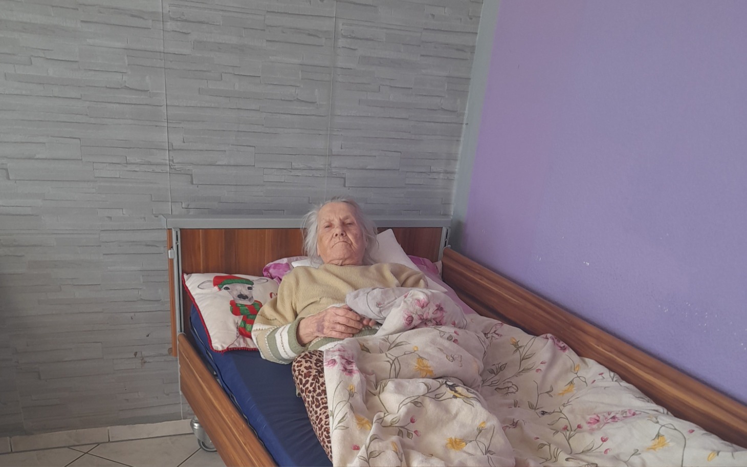 
        Starsza kobieta leży w łóżku, przykryta kocem.
    