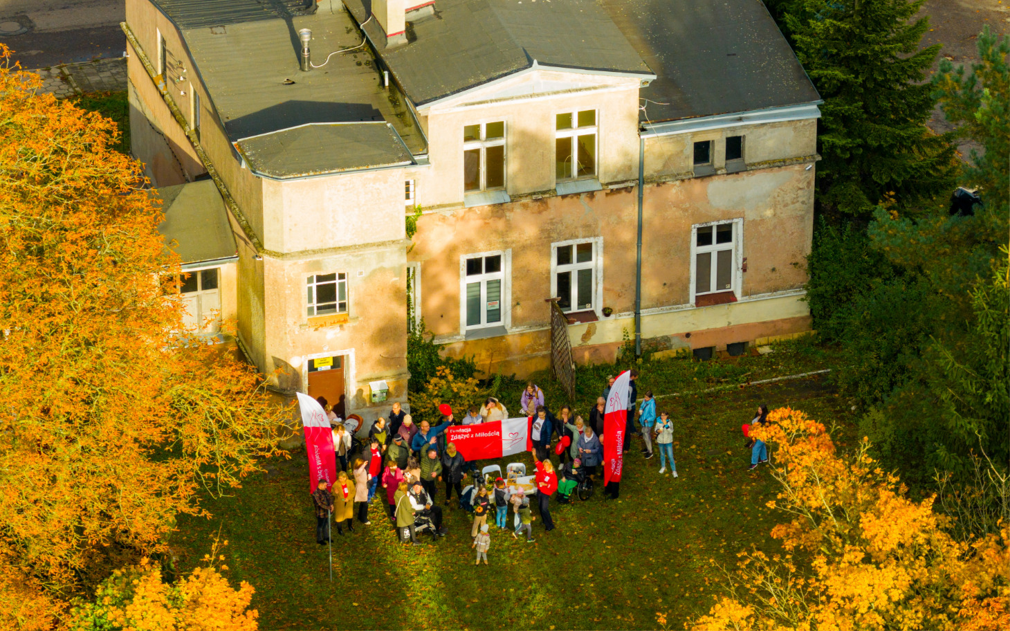 
        Grupa ludzi z flagami przed starym budynkiem otoczonym drzewami w jesiennych kolorach.
    