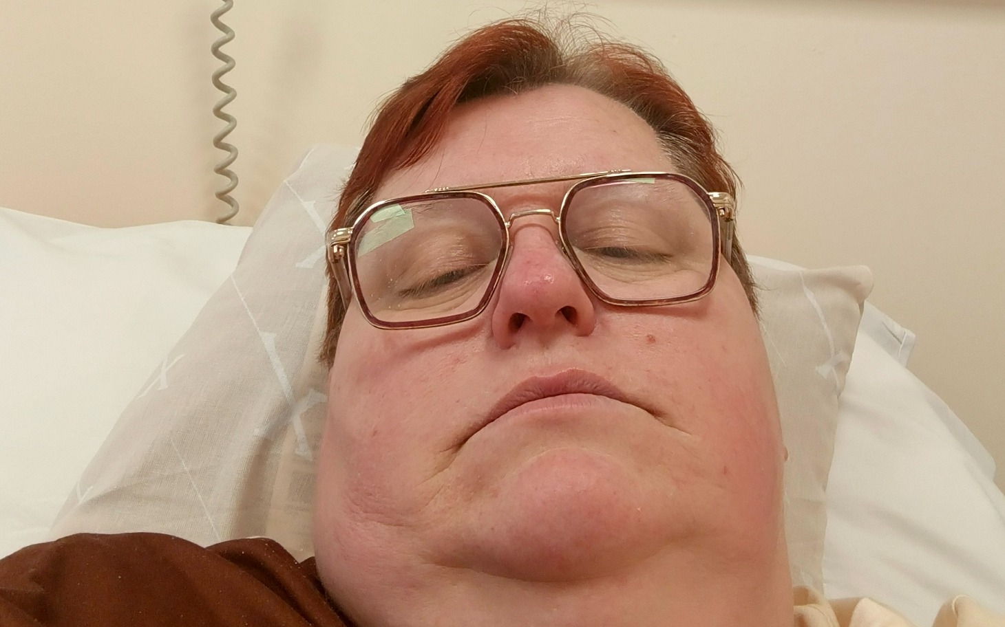 Osoba leżąca na łóżku szpitalnym, z okularami i poważnym wyrazem twarzy.